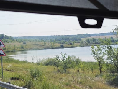 The image captures a scenic view of a rural landscape, possibly in the city of Berezki, Russia. In the foreground, a metal guardrail runs along the edge of what appears to be a road. Two triangular road signs are visible on a pole; one depicts a crossroads, and the other indicates a steep downhill slope. Beyond the guardrail, a body of water, likely a lake or river, stretches across the middle ground. The water reflects the hazy, pale sky. The banks of the water body are lined with lush green vegetation, including tall grasses and scattered trees. In the background, a dense forest covers rolling hills under a light sky, with several power lines visible in the distance. The overall impression is one of natural tranquility and open space. No people or animals are present in the scene.