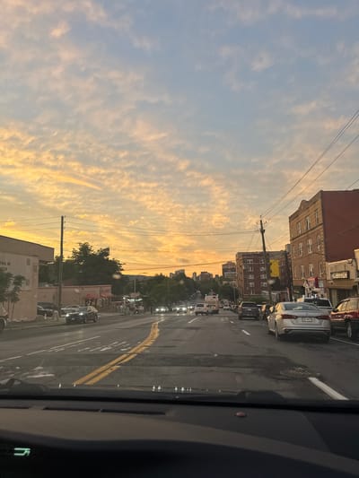 The image depicts a street view in the City of Yonkers, United States of America, during what appears to be sunset or sunrise. The sky is filled with a mix of blue and warm orange and yellow clouds. The road is wet, suggesting recent rain, and features double yellow lines marking the center lane. Several cars are visible on the road, including a silver sedan in the foreground, a dark SUV, and a red SUV further down the street. Buildings line the right side of the street, some appearing to be multi-story with brick facades. Trees are present on both sides of the road, and utility poles with wires are prominent. The perspective is from inside a vehicle, looking forward through the windshield. The overall scene conveys a sense of urban traffic during a colorful sky event.