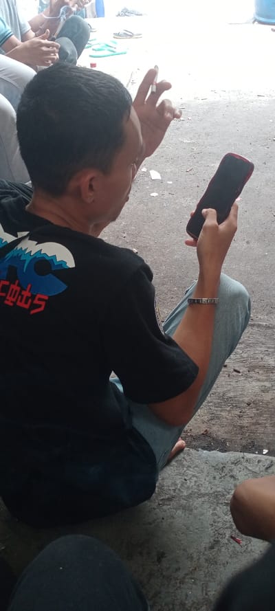 An image taken in Cilampeni, Indonesia, depicts a group of individuals seated on a rough, grey concrete surface. The primary subject is a male, viewed from behind and slightly to his right. He wears a black short-sleeved t-shirt with a white and blue wave graphic above red characters, light blue jeans, and a dark studded bracelet on his left wrist. He holds a dark-colored smartphone with a red case in his right hand, actively engaging with the screen, and a lit cigarette between the index and middle fingers of his left hand. To his left, another individual is partially visible, wearing light-colored pants and a light blue shirt, with their hands clasped. In the immediate foreground, the knees of a third individual are visible. In the background, on the concrete, light blue and dark flip-flops are visible, along with a red bottle cap and scattered debris. A blue barrel is partially visible in the upper right background.