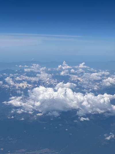 The image is an aerial view of mountainous terrain, likely captured from an airplane. The sky above is a clear, deep blue with some faint, high-altitude cirrus clouds. Below the sky, a layer of fluffy white cumulus clouds partially obscures a mountain range. The mountains themselves are rugged and appear to be snow-capped in places, indicating a high altitude. The lower slopes of the mountains are covered in dark green vegetation, suggesting forests. In the distance, there are hazy, layered mountain ranges. The image does not depict any people or specific objects within the landscape, nor does it provide enough detail to definitively locate it within Viehhausen, Austria. The scene is a natural, atmospheric landscape.