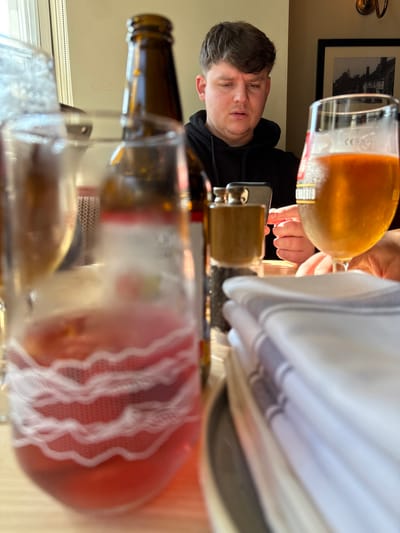The image displays an indoor scene within a dining establishment in Whiteparish, United Kingdom. The perspective is low, looking across a table.
A Caucasian male, with short dark brown hair, is seated in the mid-ground, wearing a black hooded sweatshirt. He is focused on a dark-colored smartphone, held vertically on the table directly in front of him, and his right hand is visible, interacting with its screen. His expression appears concentrated, with slightly furrowed brows and a parted mouth. Part of a second person's hand is visible on the far right of the table.
On the table in the foreground, from left to right, are: a blurred glass containing a red-pink beverage with white wavy patterns; an out-of-focus amber glass bottle with a red and white label; a sharply focused dark pepper grinder; and a stack of white, folded napkins with grey stripes resting on a grey plate. To the right of the pepper grinder, a clear beer glass, containing a golden amber liquid and displaying condensation, features "MADRID" and a "CE 0,4L" mark.
The background shows a light-colored wall. A window with vertical blinds is visible on the left, admitting bright natural light. On the right, a dark-framed picture depicting an old building or street scene hangs on the wall, illuminated by a brass-colored wall light fixture above it. The table surface appears to be a light wood tone.
