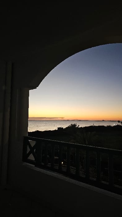 Captured from within a dark, arched structure, the image reveals a vibrant sunset over a body of water. The horizon is marked by a transition from light yellow to orange. A dark silhouette of trees and buildings line the shore. A wooden railing is visible on the right, juxtaposing the natural scene with a man-made structure. The light of the sunset reflects off the water's surface, creating a shimmering effect.