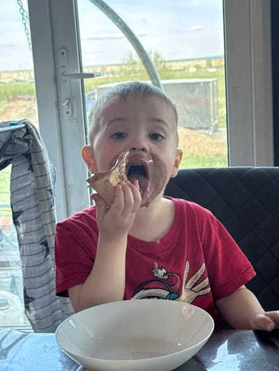A young child with chocolate smeared on their face is eating an ice cream cone at a table in a room. The child is wearing a red t-shirt with a cartoon graphic. A white bowl is near the child. A dark chair is behind the child. A window is visible to the left, showing greenery and an overcast sky. A grey and white patterned cloth is beneath the table near the window. The precise location in the city of Тоншалово, Russia, cannot be determined from the image.