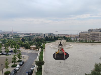 An elevated, wide-angle, daytime shot captures a city skyline with a body of water and a playground. The sky is overcast with a light gray hue. In the foreground, a rooftop patio paved with light gray cobblestones features a children's playground with a red slide, a yellow swing set, and a small, dark-roofed playhouse. A white pergola structure is visible behind the playground. The patio area is bordered by a low white wall.
To the left of the patio, a multi-lane road curves downwards, lined with parked cars. Several trees are planted along the sidewalk and median. Beyond the road, a park-like area with more trees leads to a town with residential buildings and industrial structures in the distance. Prominent features in the middle ground include a tall, red and white striped smokestack, various buildings, and what appear to be port facilities along the water. A large, ornate, light-colored building with many windows and a dark mansard roof dominates the right side of the frame, appearing to be an apartment building or hotel.
The distant background shows the expanse of a large body of water, likely the Caspian Sea, with several cargo ships and possibly oil rigs visible on the horizon. The overall impression is of a mixed urban and industrial landscape with a residential area and recreational space. The location is identified as Khatay Raion, Azerbaijan.