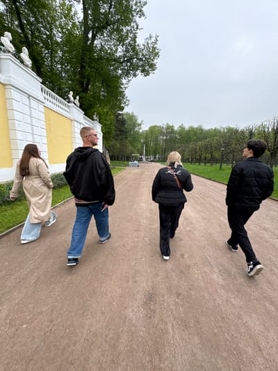 Four individuals are observed on a pathway within the State Museum Estate "Arkhangel'skoe" in Russia. The pathway, which is a reddish-brown color, appears to be lined with green grass on either side. The first individual is male, dressed in a black puffer jacket and black pants, and is walking away from the camera. The second individual, a female with blonde hair, is wearing a black puffer jacket with a brown strap and black pants and is also walking away from the camera. The third individual, a bald male wearing glasses, is wearing a black jacket and blue jeans. The fourth individual, a female with long brown hair, is wearing a tan trench coat and light blue jeans. A yellow and white wall is visible to the left, with trees lining the background. The sky appears overcast.