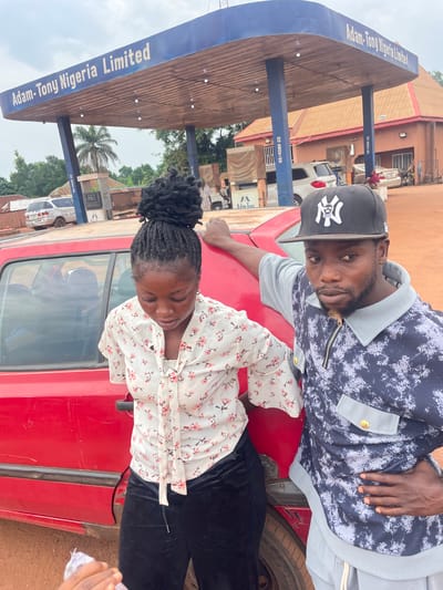 A man and a woman are posing in front of a red car at an Adam-Tony Nigeria Limited gas station in Otukpa, Nigeria. The man is wearing a baseball cap and a patterned shirt, and he is leaning on the car. The woman has black braids and is wearing a white shirt with pink flowers and black pants; she is standing in front of the vehicle. A gas station canopy is visible in the background, along with other buildings and vehicles.