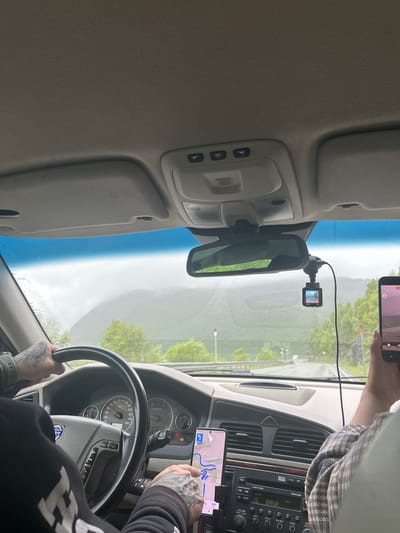 The media file captures an interior view from the driver's seat of a car. A person's hands, adorned with tattoos, are visible on the steering wheel and operating a smartphone mounted on the dashboard. The phone displays a navigation app with a map and route. A dashcam is affixed to the windshield, and a passenger's hand holding another phone is also visible, showing a video feed from a recording device. The car's dashboard, including the instrument cluster and radio, is partially visible. Outside the car, through the windshield, a green, rural landscape with trees and a road can be seen. The sky appears overcast, suggesting potentially rainy or foggy weather. The lighting indicates it is daytime. The overall scene conveys a sense of travel or a road trip.

Visible text: "ERROR" on the car's radio display, "VOLVO" on the steering wheel, and navigation details on the smartphone. The location is Røldal, Norway.