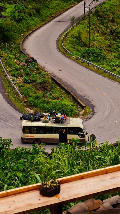 A medium shot photograph shows a winding road in Phố Bảng Commune, Vietnam. A beige passenger van, its roof laden with luggage and a motorcycle, is paused on the road. The van is marked with the number "23B-001.67". A person stands in the open doorway of the van. The road winds through green hills, and a metal guardrail runs along part of it. In the foreground, a wooden surface with a plant pot is visible, suggesting the photo was taken from an elevated position.