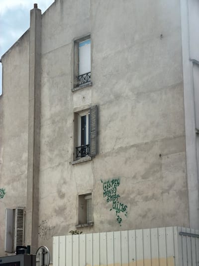 The image shows the exterior of a concrete building, viewed from below. The concrete is rough and shows signs of wear, with some cracks visible. There are three small windows or vents on the surface. The left window is open, revealing a white ceiling and some metal bars. The middle window is also open, with a view of a dark interior and a decorative metal grille. The right window is smaller and appears to be a vent.
Green graffiti is visible on the concrete. One piece of graffiti reads "GIS P EH MONTO SMET" and another reads "MI MS . 23.". There are other faint markings on the wall.
To the right of the concrete wall is a section of corrugated metal siding, painted in alternating bands of white and pale yellow.
The overall impression is of an urban or industrial setting, possibly a less-maintained part of a building. The sky is visible in the lower left corner, suggesting it's daytime, but the lighting is somewhat muted, and the clouds are not clearly discernible. The perspective is unusual, looking upwards at the underside of a structure.