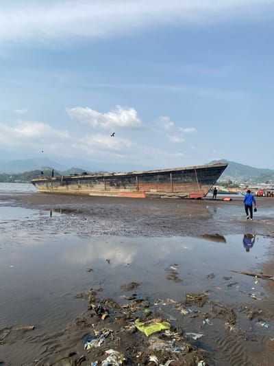 The image displays a bright daytime scene along a coastal area, likely in Victoria, Cameroon, with a large, long wooden boat beached on the wet sand. The boat, appearing dark and weathered, stretches horizontally across the middle ground, indicating a possible low tide. Its plank construction is visible along its side.
Two main figures are near the boat. A person in a vivid blue shirt and dark trousers stands prominently near the boat's stern, their reflection clearly visible in the shallow water beside them. Another smaller, darker figure is positioned closer to the boat's midsection. Further up the shore in the background, more indistinct figures can be seen.
The sky is a clear, light blue with scattered white clouds, some of which are reflected in the tranquil water. Beyond the coastline, a backdrop of green, tree-covered hills or mountains rises in the distance. A significant detail is the extensive accumulation of trash and debris, including plastic items and other refuse, scattered across the darker, drier sand on the right side of the image. There is no visible text. The scene depicts a working coastal environment, characterized by traditional watercraft and visible environmental pollution.