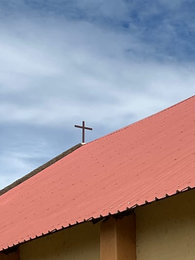 A close-up, low-angle outdoor shot captures a cross atop the red, corrugated metal roof of a building. The cross is dark brown and centrally positioned on the peak of the roof. The roof itself is angled diagonally from the bottom left to the top right of the frame, with visible fasteners dotting its surface. The building's exterior wall, made of light brown textured material, is partially visible at the bottom. Above the roof, the sky is a pale blue with scattered, wispy white clouds. The scene is static, with no people or discernible activity. The image is well-lit, suggesting daytime. The location is identified as Bawan Dodo, Nigeria.
