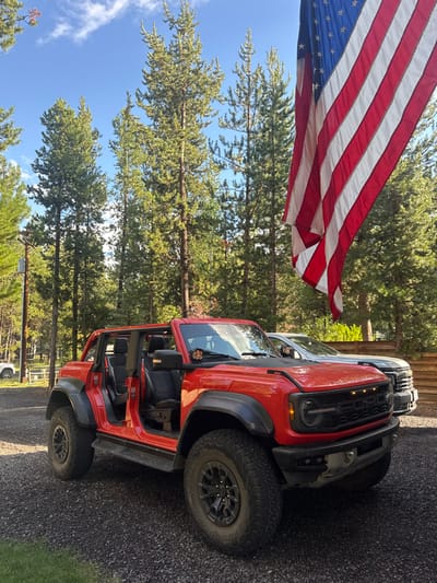A red four-door Ford Bronco Raptor, with its doors removed, is parked on a dark gravel driveway. The vehicle displays large off-road tires, black wheels, and cowl-mounted auxiliary lights. Behind it, a light-colored pickup truck, identified as a Chevrolet Silverado HD, is parked near a wooden fence or building. A large American flag hangs vertically from the upper right, partially obscuring the background. The scene is set outdoors under a clear blue sky with some white clouds, featuring numerous tall coniferous trees, predominantly Ponderosa Pines, indicating a forested or semi-forested residential area. A utility pole is visible on the left. The environment and tree species are consistent with the natural landscape of Bend, Oregon, United States.