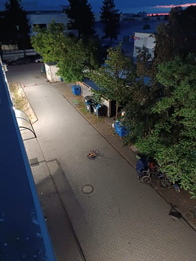 This is a high-angle, eye-level, medium shot taken at dusk or dawn in Germering, Germany, likely an industrial or commercial area. The scene depicts a paved area with interlocking brickwork. On the left, a blue wall of a building forms the edge of the frame. Adjacent to the building, a covered walkway with a translucent roof extends across the paving stones. Further along the walkway, a small, white, portable storage unit with a ladder leaning against it is visible, next to a blue dumpster.
Dominating the right side of the image are large, leafy green trees. Scattered throughout the scene are several large dumpsters, some blue and some black. Paved pathways meander through the area. In the middle of the paved path, a small, red and yellow children's bicycle is lying on its side. Further down the path, near the trees, a group of bicycles are parked, some covered with a blue tarp. One bicycle in this group is a child's bike with blue and yellow accents. A dark-colored, rectangular object, possibly a laptop or briefcase, lies on the ground near the parked bicycles. In the distance, several buildings are visible, including one with a sign that reads "PROmetalt". The sky exhibits the faint colors of twilight, with a reddish-orange hue near the horizon. No people are clearly visible in the image.