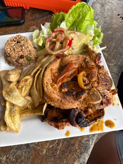 A white rectangular plate of food is centered on a textured, dark wooden tabletop in La Ceiba, Honduras. The plate contains two grilled pork chops, one largely visible and featuring a bone, both topped with a reddish-brown sauce containing sliced bell peppers and onions. To the upper left of the plate, a scoop of brown rice is mixed with black beans and green seeds. Below the salad and next to the rice, a pile of thinly sliced, fried plantains (tajadas) is present. Above the pork chops, a small portion of green leaf lettuce salad is visible, dressed with red onion rings, tomato slices, and a creamy yellow sauce. In the background, a dark mobile phone and a partially visible orange and black object rest on the table. A human leg, clad in dark shorts, is partially visible in the lower right quadrant, suggesting a seated individual. No direct actions or interactions are depicted beyond the prepared meal.