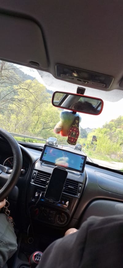 **Location:** The image appears to be taken from the inside of a vehicle located on a road, with vegetation and mountains visible through the windshield, near Krupnik, Bulgaria.

**Objects:**
*   A tablet and smartphone are mounted on the vehicle's dashboard.
*   A rear-view mirror with a rainbow-colored tassel and a tag attached to it.
*   Steering wheel and gear shift.
*   Car air vents and central console.

**People:**
*   A person is behind the steering wheel of the vehicle.

**Environment:**
*   A mountainous and green landscape is visible outside the vehicle.