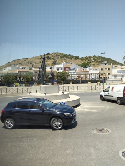 The image shows a street scene in Cabra, Spain, featuring a roundabout with a monument, vehicles, and buildings in the background. The monument appears to be a bronze sculpture depicting two figures, one holding a flag and the other a drum, placed on a circular concrete base. A dark-colored car is driving in the foreground, moving from left to right across the frame. On the right side of the roundabout, a white van is also visible, heading in the same direction. The background consists of a hillside covered with vegetation and houses, with a clear blue sky above. The architecture of the buildings suggests a Mediterranean style. The street is paved with asphalt and marked with white lines. There are streetlights visible in the background. The overall impression is that of a bright, sunny day in a Spanish town.