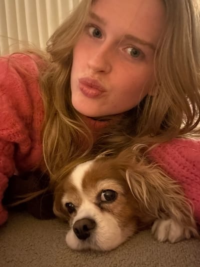 A young woman with blonde hair poses for a selfie with her Cavalier King Charles Spaniel. The woman wears a pink sweater and makes a kissing face towards the camera. The dog's head rests next to hers, with its distinctive brown and white markings and expressive eyes. They appear to be lying on a carpeted surface, with the dog partially obscured by the woman's sweater. The photo is taken at a close angle, emphasizing their faces.