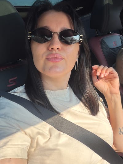 A woman with dark hair and sunglasses is sitting in the driver's seat of a car. The car's interior is visible, including the headrest with a red "S" logo and a seatbelt. The woman is wearing a light-colored t-shirt and has a tattoo on her left forearm. The lighting suggests it is daytime, possibly late afternoon. The image does not contain any information indicating a location in Bolshiye Dvory, Russia.