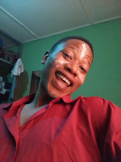 A young African man is smiling broadly, revealing his teeth. He has white markings on his forehead and cheek. He is wearing a red shirt with the collar open. The background is green, and there is a metal shelf with objects on it to the left. The location is indoors. There is no information given about the city of Oshogbo, Nigeria.