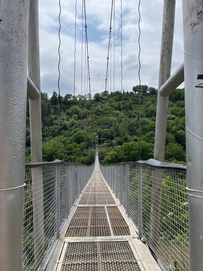 The scene depicts a frontal, eye-level view of the Khndzoresk Swinging Bridge in Khndzoresk, Armenia, extending across a deep, forested gorge. The bridge features a long, narrow walkway composed of grey metal grating panels and is flanked by high metal mesh-paneled side barriers. Two large, cylindrical, silver-grey metal support columns stand in the immediate foreground, framing the bridge's perspective. Numerous thin, wavy metal suspension cables descend vertically from unseen overhead main cables, supporting the bridge deck. The surrounding environment consists of steep hills densely covered with vibrant green deciduous and coniferous trees. The sky above is largely overcast with discernible white and grey cloud formations. No individuals, animals, or specific activities are observed on the bridge or within the visible landscape.