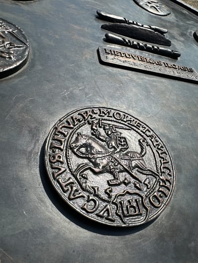 The image shows a close-up of a bronze relief on a dark, textured surface, likely part of an outdoor monument or information board. In the foreground, a circular bronze medallion features a detailed relief of a knight on horseback, a symbol commonly associated with Lithuania, specifically the "Vytis" or "Pahonia" motif. The knight is depicted with a sword raised, and a shield bearing an emblem. The medallion is surrounded by text in an archaic script, partially obscured.
In the middle ground, a rectangular bronze plaque is visible with raised text that reads "LIETUVISKAS ILGASIS" followed by what appears to be Braille. Above this plaque are three horizontal, elongated bronze elements, resembling stylized representations of traditional Lithuanian tools or weapons.
In the background, another circular bronze element with relief details is partially visible, suggesting this is part of a larger artistic installation. The lighting indicates an outdoor setting with strong sunlight casting shadows.
This monument is located in the city of Trakai, Lithuania, specifically at the Trakai Island Castle complex. The reliefs likely serve to inform visitors about Lithuanian history, culture, and symbols.