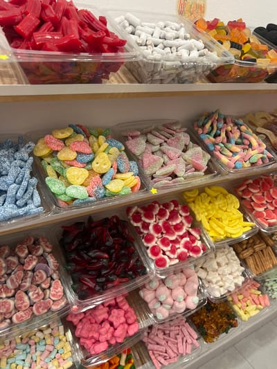The image shows a close-up, eye-level view of a candy display shelf in a store. The shelf is stocked with numerous clear plastic containers filled with a variety of colorful candies. The top shelf holds containers of red licorice sticks, white chalky candy sticks, and various gummy candies in orange, yellow, red, and black. The middle shelf displays blue sour straws, sour gummies in green, yellow, and red, pink and white sour heart-shaped candies, and multicolored gummy worms. The bottom shelf contains red gummy sharks, pink gummy babies, and pink gummy stars. There are also containers of yellow banana-shaped gummies, white gummy stars, and golden brown churro-like candies. In the lower right corner, there are more gummies and candy sticks. The background is a white wall, and the lighting appears to be bright and even, suggesting an indoor commercial setting. There are no people visible in the image. Based on external information about the location, this candy store is situated in Port de Pollença, Spain.