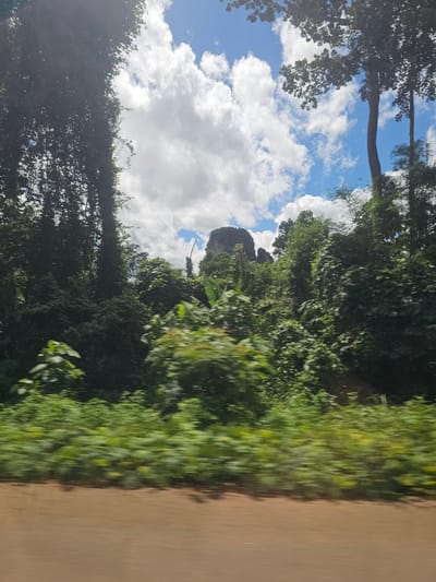 This is a landscape image taken from a vehicle traveling on a dirt road. The scene is dominated by lush green foliage, indicating a tropical or subtropical environment, consistent with the location context of Ban Nai Sa, Thailand. Dense trees and vegetation line the left side of the road, obscuring much of the view beyond. In the distance, a distinct rock formation with a pointed peak rises above the trees, a prominent landmark in the landscape. The sky is a vibrant blue, dotted with fluffy white clouds, suggesting fair weather, likely during daylight hours. The right side of the image shows the textured surface of a dirt or unpaved road, with motion blur indicating the movement of the camera. The overall impression is one of natural beauty and a journey through a verdant, possibly mountainous, region. There is no text visible in the image.