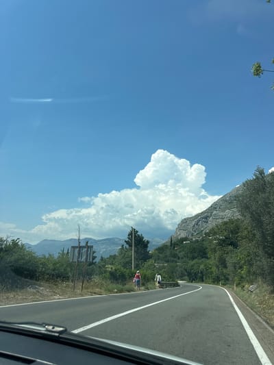 The image depicts a scenic view from a moving vehicle along a winding road. The sky is a brilliant blue, with large, fluffy white cumulus clouds dominating the upper left portion of the frame. A rugged, rocky mountainside covered in lush green trees rises to the right, bordering the road. The road itself is asphalt, with a clear white line marking its edge, curving into the distance.

In the middle ground, a few figures are visible on the side of the road. One person appears to be standing, wearing a red top and blue shorts. Another figure, possibly also standing, is partially obscured. There are also some signs along the roadside.

The scene suggests a bright, sunny day, likely midday, given the clear skies and the quality of light. The mountainous landscape and the winding road hint at a location in a hilly or coastal region. The presence of vegetation indicates a temperate climate. The overall impression is one of a pleasant journey through a beautiful natural setting. There is no visible text in the image.