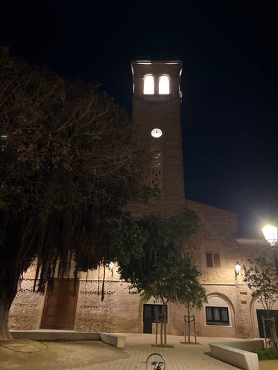 This nocturnal image captures a historical stone building, likely a church or tower, under a dark sky in La Torre, Spain. The central feature is a tall, sturdy stone bell tower, illuminated from within its upper section where two bright, rectangular openings suggest the presence of bells. Below these openings, a white circular clock face with black hands clearly indicates the time as approximately 10:10.

In the foreground and midground, a variety of trees stand. Some appear bare-branched and skeletal against the dark night, while others retain denser green foliage. One tree in the lower right seems to bear small, round fruit, possibly lemons or oranges.

To the right of the tower, a lower section of the same or an adjacent stone building is visible. This section features arched doorways or windows, a window with wooden shutters, and another with a dark grill. Overhead, a bright street lamp casts a warm, yellow glow onto the stone facade and the paved ground below. The paving stones are light-colored, suggesting a public square or pathway. A modern-looking concrete bench or low wall is positioned along the right edge of the frame, and a metal bicycle rack is partially visible at the bottom right.

The overall atmosphere is tranquil and historical, with the artificial lights creating strong contrasts between the illuminated architectural details and the deep shadows of the night. No human activity is visible, contributing to the peaceful ambiance of the scene. The weather appears clear. No explicit text is readable beyond the numbers on the clock face.