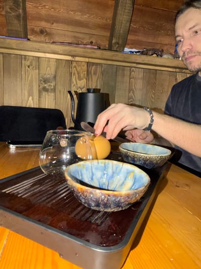 A man is seen preparing an egg in a wooden room. A clear glass pitcher holds a yellow egg, balanced with a spoon. Two decorative bowls sit nearby on a glossy surface. A black kettle rests on the table. The room features rustic wood paneling on the walls and floor, creating a cozy, warm ambiance.