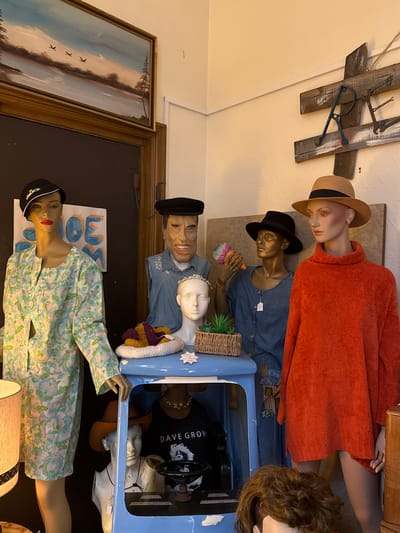 The image shows a collection of mannequins in what appears to be a shop or display setting. From the bottom, there's a mannequin wearing a green and blue floral dress and a black cap, positioned near a lamp. Above it is a blue toy car containing multiple mannequins, one wearing a "Dave Grohl" shirt. Next to the car is a mannequin with a denim shirt and a black hat, a blonde wig and a small white mannequin wearing a bridal tiara. Another mannequin in denim holding a prop that looks like an ice cream. Behind these, there's a mannequin in an orange sweater wearing a straw hat. In the background, there is a wooden airplane decoration on the wall and a framed picture on the left. The photo was taken at The Margate Caves in Thanet, United Kingdom.