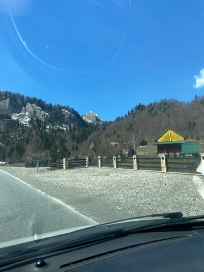 The image depicts a mountainous landscape in Sedlačka gora, Montenegro, as viewed from inside a vehicle. The sky is clear blue with a bright halo. A mountain range rises to the left, partially covered with snow and sparse vegetation. To the right of the mountain, a structure with a yellow and green roof stands behind a fenced area. A grey asphalt road runs alongside the fence. The lower right corner of the image features the interior of a car, including the windshield and parts of the dashboard.