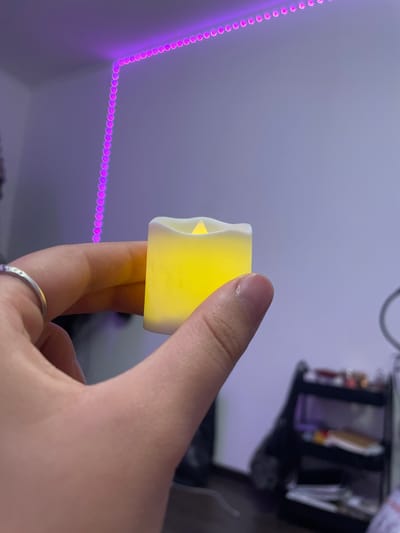A hand holds a small, battery-operated candle with a yellow LED flame. The candle is positioned in the foreground of the image, and the hand is partially obscuring it. In the background, a string of purple LED lights runs along the top corner of a room. The room appears to be a bedroom, with a shelf unit visible to the right, filled with various items. The overall lighting of the scene is dim, with the exception of the bright LED candle and the purple lights. There is no indication of the location being Borisova Griva, Russia, or any other specific city.