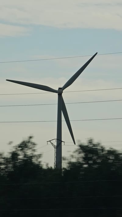 An outdoor, low-angle, vertical, medium shot shows a wind turbine silhouetted against a pale, overcast sky. The turbine's three blades are angled upward and to the left and right. The turbine's tall, slender tower extends from the bottom of the frame. Three parallel power lines run horizontally across the frame, intersecting the tower. In the foreground, a blurred, dark green line of trees or foliage stretches across the lower half of the image. A blurred, metallic pylon supporting electrical wires is visible on the right side of the turbine tower. The motion blur suggests the image was taken from a moving vehicle. The location is identified as La Muela, Spain.
