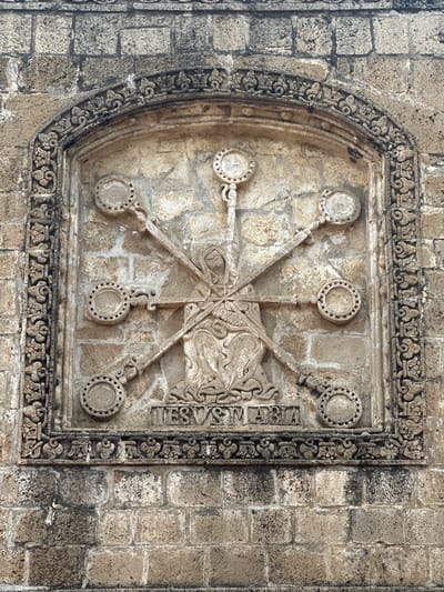 The image displays a relief carving mounted on a stone wall in San Andrés Calpan, Mexico. The carving, framed by an ornate border, depicts a seated figure, possibly a saint or religious icon, holding an infant. Radiating outwards from the figure are eight ornate, circular objects, each suspended from a rod. Below the central carving, the inscription "IESVS MARIA" is prominently displayed. The surrounding wall is constructed of rough-hewn stone blocks.