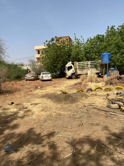 The image shows a scene in a dry, open area in Khartoum, Sudan, at coordinates 15.552 latitude, 32.57 longitude. There are two white cars parked on a dirt lot, one facing the viewer and the other facing away. Behind them is a large, white truck with a flatbed, parked on the right edge of the lot. The truck is facing the viewer and a blue water tank is visible in the background on the right, behind a cluster of trees. There is a small, two-story building in the background, with a roof that appears to be partially obscured by trees. The lot is sparsely vegetated, with a mixture of dry grass, dry leaves and sticks.  The ground is covered in dirt and dust, with some rocks and debris scattered throughout.  There are several tires lying on the ground, some arranged in a semi-circle, in the foreground. The scene appears to have been captured on a sunny day. No people are visible in the image.