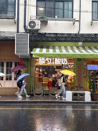 This image captures a street scene in Xuhui District, China, during what appears to be a rainy day. The setting is in front of a store with a green and white striped awning. The store's signage, visible in neon and illuminated lettering, reads "poooon!" and "舔勺!酸奶" which translates to "Licking Spoon! Yogurt." Inside the store, there are shelves stocked with items, a blackboard menu listing "Coffee, Soda, Smoothies, Waffles, Ice Band," and some decorative plants.
Several people are present, some sheltering under umbrellas. In the foreground, two individuals are walking with umbrellas, one a light blue umbrella and the other a dark red one. Further back, a person sits outside the store under a bright yellow umbrella, with a backpack beside them. Another person, leaning against the store's facade, is also visible. The ground is wet and reflective, indicating ongoing rain. There's also an air conditioning unit mounted on the exterior of the building to the left of the store. The overall mood seems calm despite the rain, with people going about their day. The time of day is not clearly discernible, but the lighting suggests it could be daytime.
