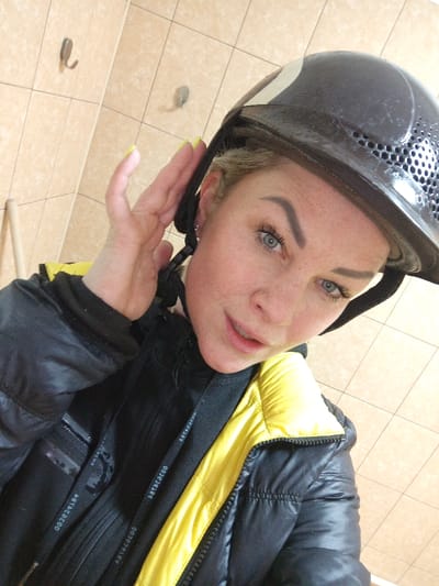 A female individual is depicted in a close-up selfie, captured at a high angle in an indoor setting. She wears a dark helmet with a white stripe and perforated ventilation. Her right hand, featuring yellow-painted fingernails, is positioned at the helmet's side, appearing to adjust it. Her face shows blue eyes, long eyelashes, defined eyebrows, and freckles across fair skin, with her mouth slightly parted. She is dressed in a black and yellow puffer jacket, zipped, over a dark garment with a visible black strap bearing the text "002829100" and "ABTACADO". The background reveals a light beige tiled wall with two metallic wall hooks and a light-colored wooden dowel visible vertically in the lower-left corner. This scene is located in the city of Gorka, Russia.