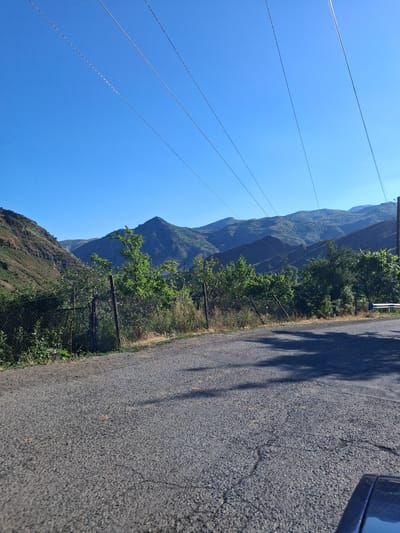This image depicts a scenic mountain landscape in Shatin, Armenia. The foreground features a paved road with shadows cast by trees, suggesting it is daytime. Power lines crisscross the bright blue sky, indicating the presence of infrastructure in this mountainous region. To the left of the road, a fence separates the road from a vegetated hillside. Further back, rolling green mountains rise, some with visible terraces, hinting at agricultural activity or past human intervention. Lush green trees dot the landscape, especially near the fence line. The clear sky and bright sunlight suggest fair weather, likely mid-morning or afternoon given the shadow lengths. The overall impression is one of rural beauty and tranquility, typical of mountainous terrain in the region. There is no visible text in the image.