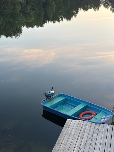 The image shows a blue rowboat with a small outboard motor attached to the stern, docked at a wooden pier. A red life preserver is visible inside the boat. The boat is on dark water, and the reflection of trees and the sky is visible on the water's surface. The pier is made of weathered wooden planks. The scene appears to be set in a natural environment, possibly a lake or river, during daylight. The city of Kamysh, Russia, is not depicted in this image.