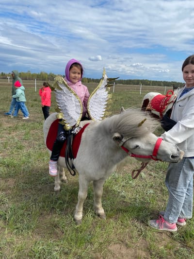 The image depicts a young girl sitting on a white pony with decorative wings attached to its back, likely in a rural area near Kumakskoe, Russia. The pony has a red saddle and bridle. A woman in a white jacket and gray pants is holding the reins of a second pony, which is piebald, standing beside the white pony. Other individuals, including one wearing a pink jacket, are visible in the background. The setting appears to be a grassy field under a partly cloudy sky.