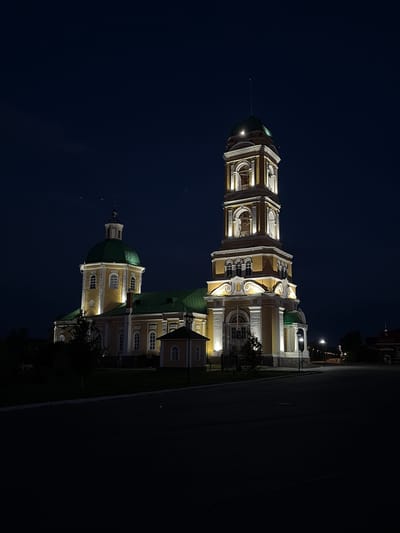 The image displays a grand architectural complex, likely a church or cathedral, illuminated at night in Nikolo-Beryozovka, Russia. The prominent structure features cream or yellow-colored walls with white decorative elements and distinctive green domes and roofs.
On the left side, a tall, ornate bell tower rises, characterized by multiple levels of arched openings, balustrades, and intricate detailing, topped with a green dome and a slender spire. To its right, the main body of the building is visible, also adorned with numerous arched windows and topped by a larger green dome. The entire complex is brilliantly lit by artificial lights, highlighting its baroque or classical architectural style against the dark backdrop.
The time of day is clearly night, with the sky appearing as a deep, clear blue-black, indicating fair weather. Several streetlights are visible in the background on the right, suggesting the presence of a road or town square adjacent to the building. The foreground consists of a dark, expansive area, possibly a paved surface or an open field, with some shadowy tree foliage near the base of the building. There are no people visible, and no specific event or activity is taking place; the scene conveys a sense of peaceful majesty. No text is visible within the image.