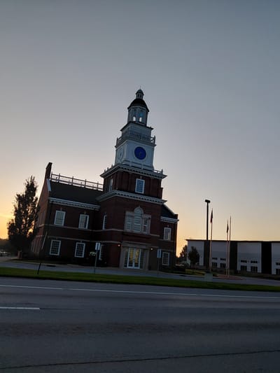 This image depicts a brick building with a clock tower, likely a government or institutional building, in Frankfort, United States of America. The building features a white clock face with a blue center, a black dome, and visible architectural details like arched windows and white trim. Adjacent to the building is a paved road, a strip of green grass, and what appears to be a sidewalk or pathway. In the background, there are additional modern buildings with large windows and flags. A tree is visible in the foreground, silhouetted against a warm, orange-hued sky, suggesting it is either sunrise or sunset. The sky is mostly clear with a gradient of light colors. The overall mood is serene and peaceful, likely captured during a quiet time of day. No people are visible in the image.