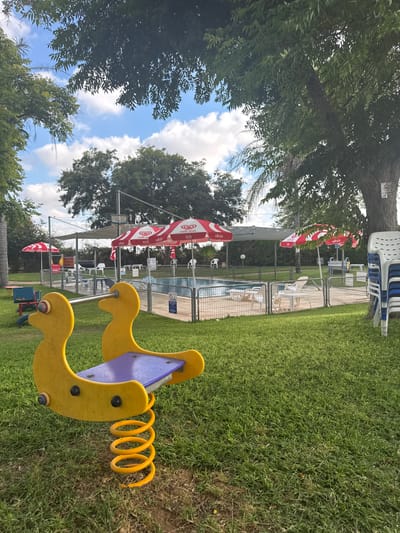 This image depicts a sunny outdoor scene at a resort or park in Beit Yitzhak-Sha'ar Hefer, Israel. In the foreground, a yellow duck-shaped spring rider for children is positioned on a green lawn. Beyond the rider, a fenced swimming pool area is visible, furnished with lounge chairs and tables, some of which are covered by red and white striped umbrellas. Further in the background, trees and foliage create a natural backdrop. The sky is partly cloudy with a clear blue expanse. A stack of white plastic chairs is situated to the right of the image. The overall impression is one of a recreational space designed for relaxation and family enjoyment, characteristic of a resort in the region.