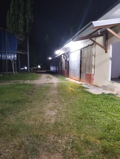 The image depicts a dirt path leading through a grassy area at night. On the right, a building with a corrugated metal roof and wooden supports is visible, with large, sliding metal doors. The building's exterior is painted in muted tones of cream and red. On the left, another structure with a blue corrugated metal facade stands. The ground is a mix of dirt and grass, with scattered debris. Trees are visible in the background against a dark sky. The lighting suggests artificial light sources illuminating the area. The location is identified as Palohgadeng, Indonesia.