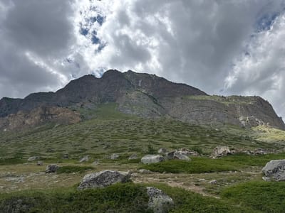 An expansive, overcast sky with dramatic, swirling clouds dominates the upper portion of the image. Below, a colossal, rugged mountain with layered rock formations rises sharply. Its slopes are a mosaic of sparse green vegetation and exposed stone. The lower foreground features a rocky, grassy terrain dotted with large boulders and patches of dense, low-lying green shrubbery. A small, scattered pile of stones, possibly the remnants of a structure, is visible on the middle right. The scene is devoid of any human presence or activity. The geographical context is El'tiubiu, Russia, implying a high-altitude, mountainous environment.