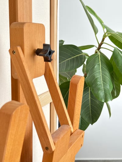This is a close-up, eye-level, medium shot of a wooden easel with a canvas stretched across it, positioned indoors. To the right of the easel, a large green ficus plant with broad leaves is visible, partially obscuring a white wall and a grey floor. The easel is made of light-colored wood, featuring adjustable arms and a black star-shaped knob used for securing the canvas. A screw is visible on one of the wooden arms. The lighting is bright, suggesting an indoor setting with ample natural light. There are no people or animals present in the image, and no specific actions are depicted. The scene is static and focuses on the details of the easel and the plant. The location is identified as Mesogi, Cyprus, though the image itself provides no explicit visual clues to confirm this specific geographical placement.