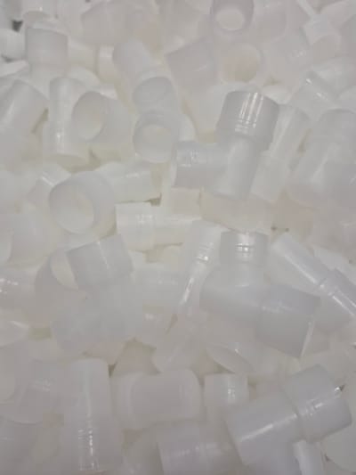 A pile of white plastic T-shaped connectors is shown in a close-up, overhead shot. The connectors are made of translucent plastic and have a smooth, cylindrical shape. They are arranged in a jumbled, disorganized manner, with some connectors overlapping others. The lighting is bright and diffuse, creating a shadowless effect. The background is a blurred mass of white, suggesting that the connectors are piled high. There are no people or any other objects visible in the image. The scene appears to be indoors, possibly in a manufacturing or storage facility. There is no information in the image to suggest a specific location in Pabradė, Lithuania.