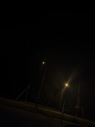 The image depicts a dark outdoor scene at night, possibly in a suburban or industrial area. Two streetlights are visible, one emitting a strong, bright light that casts rays, and another dimmer light further in the background. A small, distant bright object, possibly a star or a very distant light source, is present in the upper left quadrant of the sky.
In the foreground, a paved area, likely a sidewalk or parking lot, is visible with a patterned surface. Adjacent to this is a grassy area and a road or pathway. A metal barrier or fence runs along the edge of the road. Further back, trees and foliage create a dark, silhouetted backdrop.
On the right side of the image, a signpost with multiple signs is present. One sign is red and features white text that appears to be in Cyrillic script, with an arrow pointing to the right. Another, taller signpost to its left has vertical text that is illegible due to the darkness and angle.
There are no people visible in the image. The environment is characterized by darkness, artificial lighting from the streetlights, and vegetation. The overall impression is one of a quiet, dimly lit night scene, possibly on the outskirts of a settlement. The presence of the Cyrillic sign suggests a location where this language is used, and if it were in Lubnya, Russia, it would indicate a Russian context.