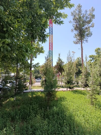 The image depicts a park-like setting in Shahrisabz City, Uzbekistan, under a clear blue sky. A tall, vibrantly colored tower, possibly part of an amusement ride or attraction, stands prominently in the background, its structure segmented into red, orange, yellow, green, and blue sections. Surrounding the tower and filling the foreground is lush, green vegetation, characterized by dense, low-lying plants and scattered trees of varying heights and species, including pine trees and deciduous trees with broad leaves.

In the mid-ground, a paved pathway is visible, bordered by manicured green spaces and some flowering plants. A white car is parked along the pathway, and a person can be seen in the lower left portion of the image, partially obscured by trees. The overall impression is of a well-maintained public space on a bright, sunny day.